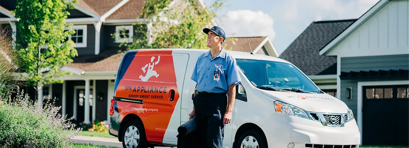 Smiling male Mr. Appliance service technician with tool bag walking away from landscaped residential home.