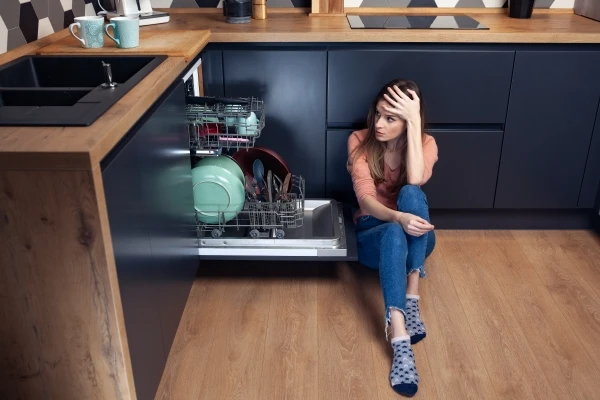 A woman sitting on the kitchen floor next to an open dishwasher, holding her head in frustration