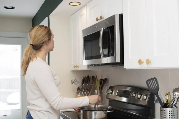 A woman looking up at a recessed light in a kitchen while stirring something on a stove