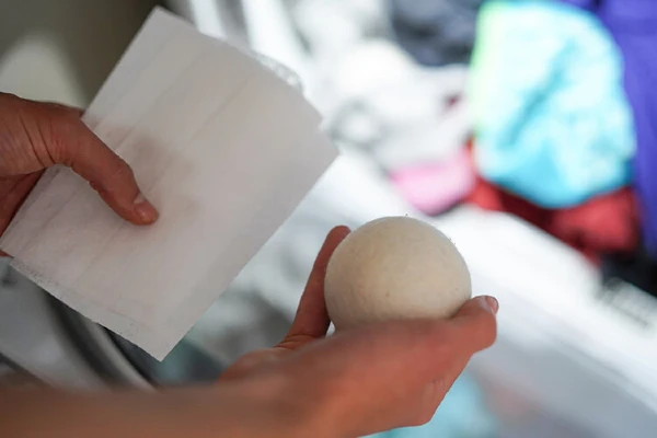 A person holding a dryer sheet and a dryer ball with laundry in the background