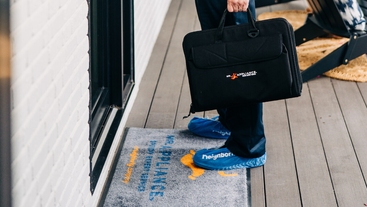 Mr. Appliance technician wearing Neighborly branded booties and carrying briefcase at front door of a home.